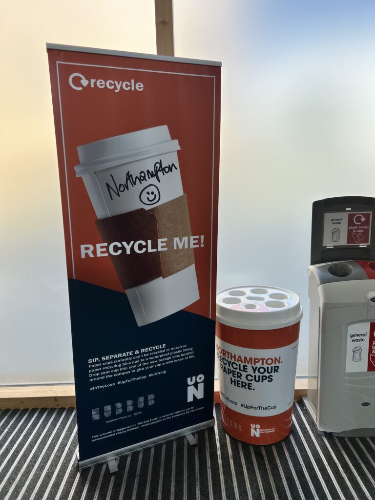 A pull up banner stands next to a specialist bin for paper cup recycling. The bin has dedicated slots for liquids, lids and paper cups.