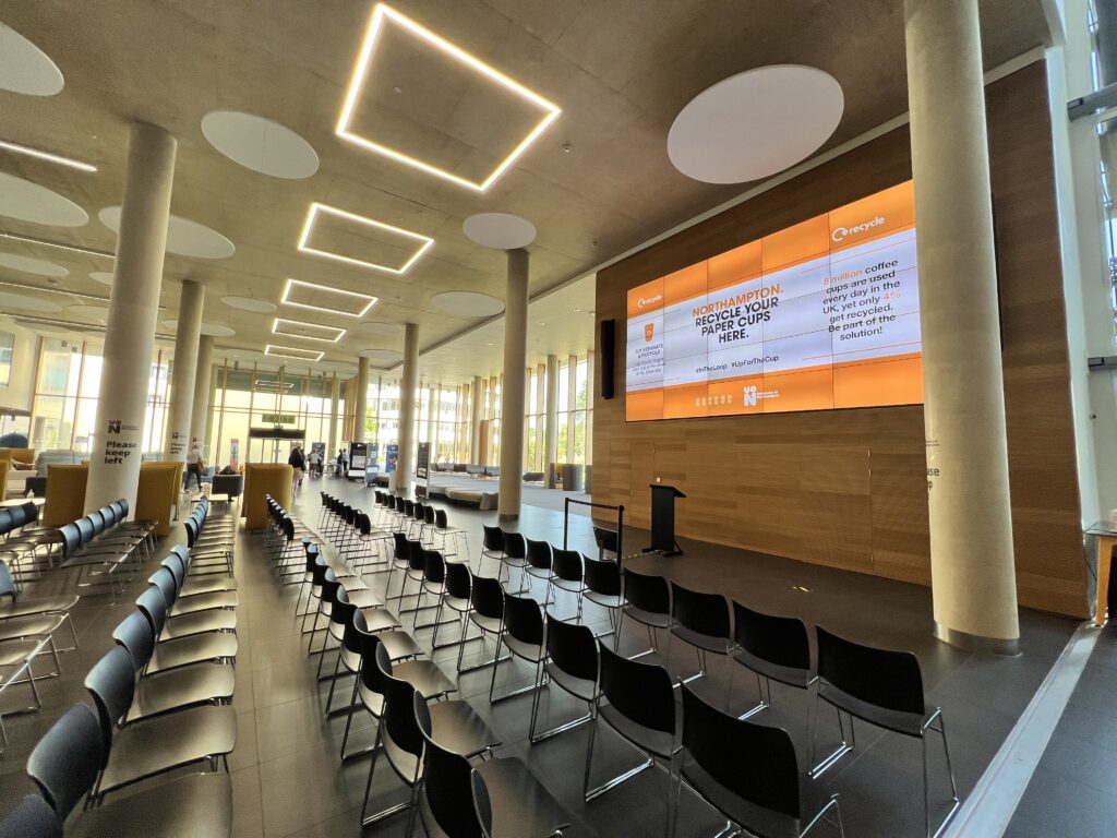 The atrium has a large screen on the wall and rows of chairs have been lined up looking towards it. A podium is set infront, ready for a presentation.