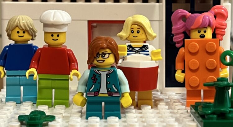 Five extremely diverse LEGO (toy) MiniFigures are standing in front of a white house with red framed windows.